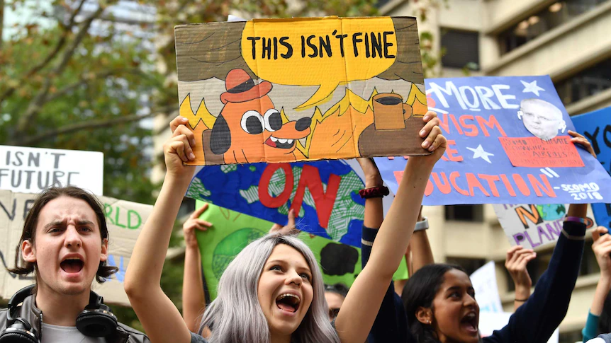 A young woman holding a sign that reads "this isn't fine", featuring a cartoon dog sitting in a burning room.
