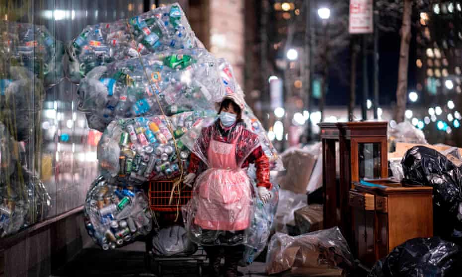 A woman wearing a face mask and a plastic bag pulls a cart loaded with bags of recyclables through the streets of Lower Manhattan.