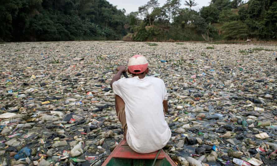 A man on the prow of a small boat paddles through a floating carpet of rubbish covering the surface of a wide river