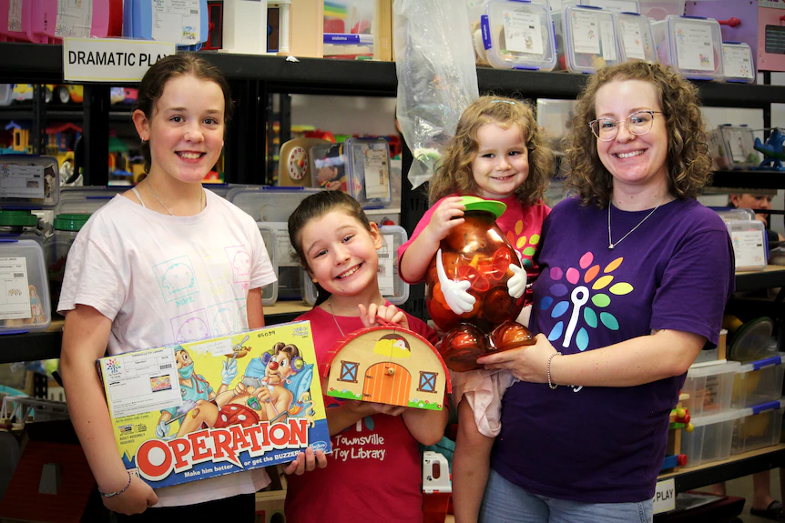A mother and her three daughters pose and smile while holding toys, in front of a shelf of more toys