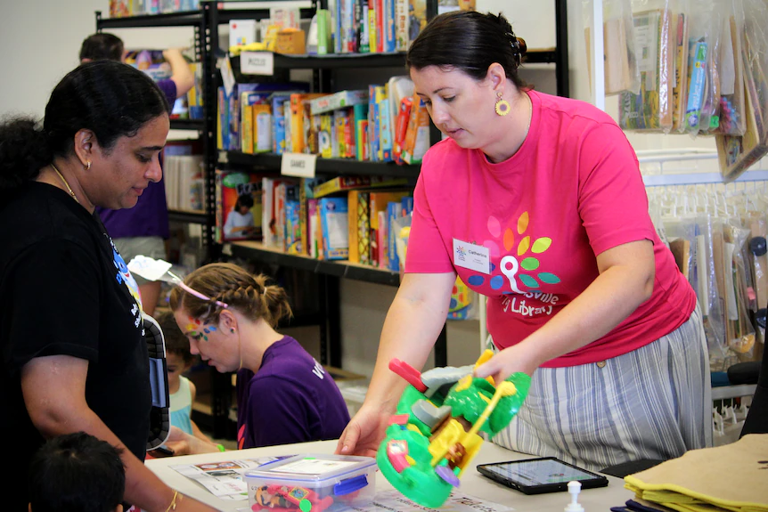 A woman in a pink shirt behind a desk serves toys to a customer 