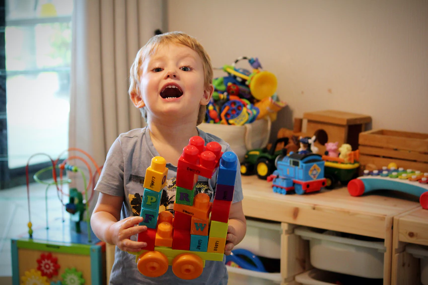 A toddler in a play room filled with toys holds a colourful truck made of blocks.