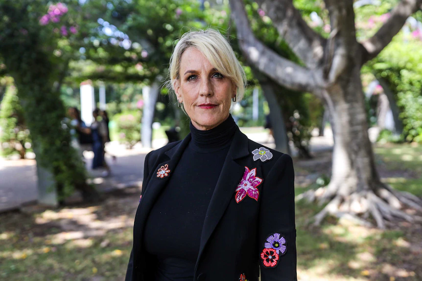 A woman with blonde hair wearing a dark suit with a floral design stands in front of some trees.