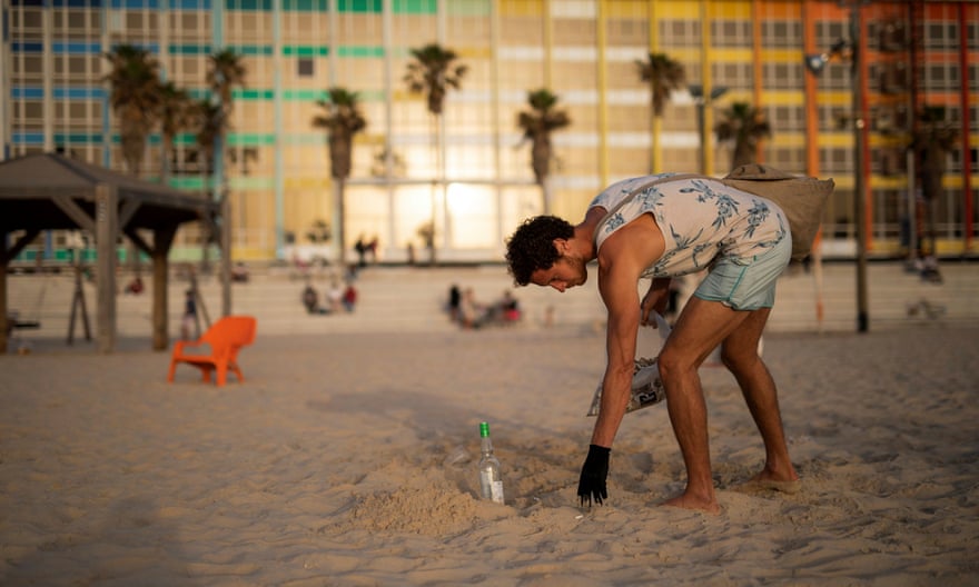 A man on a beach holding a plastic bag full of cigarette butts leans over to pick another cigarette butt from the sand. 